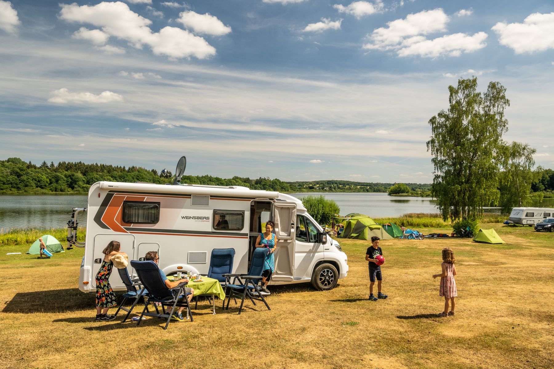 Eine Familie genießt einen sonnigen Tag neben einem Wohnmobil, das in der Nähe eines Sees geparkt ist, mit Zelten, Liegestühlen und einem Picknicktisch, der auf einer Wiese aufgestellt ist. Kinder und Erwachsene sind im Freien, umgeben von Bäumen und blauem Himmel.
